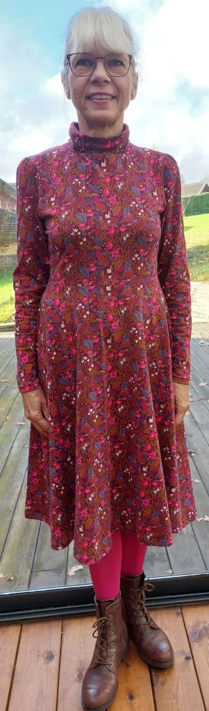 En kvinna i en blommig klänning med hög hals, står på en trätrappa. Hon bär även rosa leggings och bruna kängor.