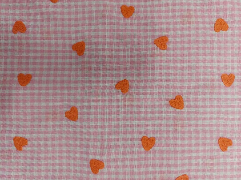 En rosa rutigt tyg med broderade orange hjärtan utspridda över ytan.